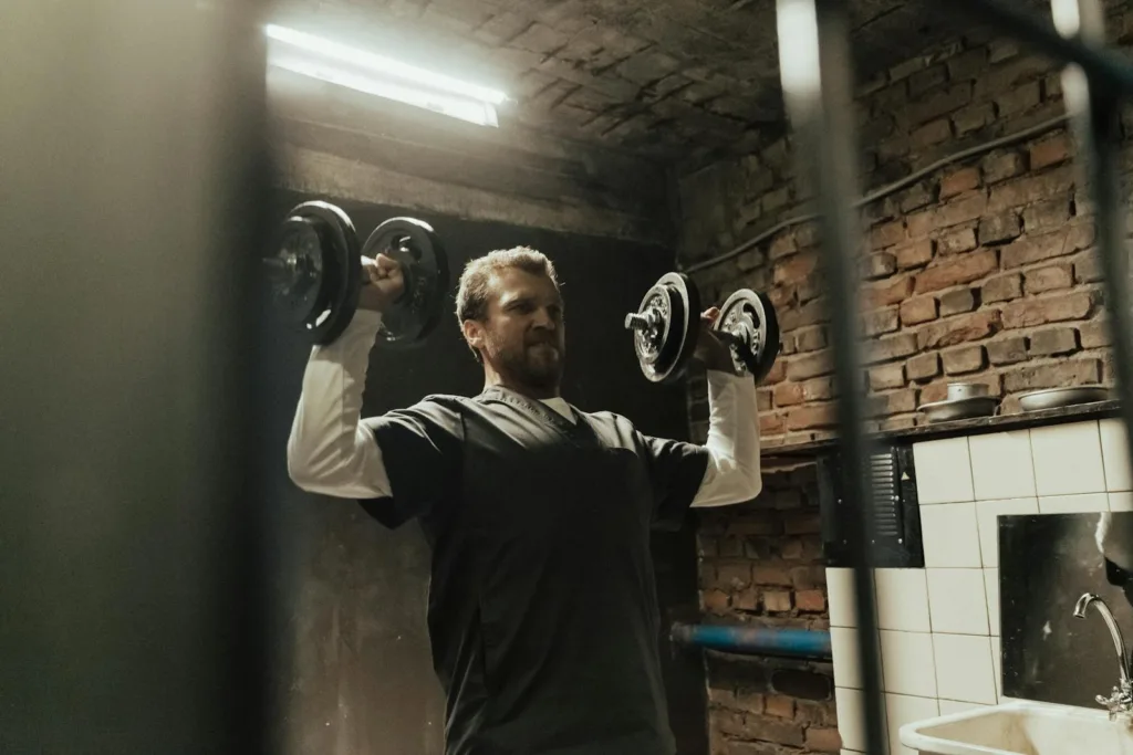 An American man exercising with dumbbells as part of full body workouts at home for beginners in a dimly lit industrial setting